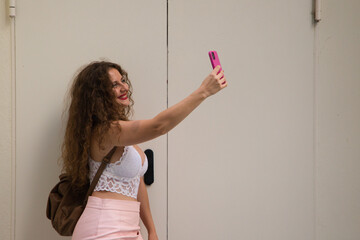Beautiful woman, young and blonde with curly hair and blue eyes is taking a picture with her cell phone and is happy. The woman is wearing a short pink skirt and white top.