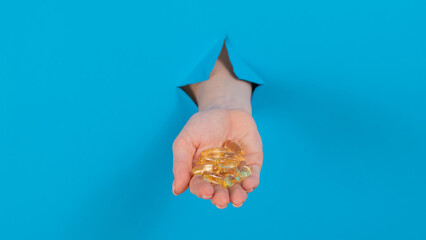 A woman holds a handful of omega 3 pills. A woman's hand sticks out of a hole in a cardboard blue background.