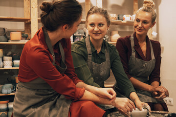 Three female friends smiling and talking while working on potters wheel making clay handmade craft in pottery workshop