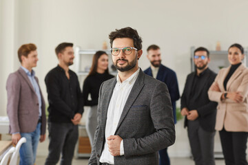 Portrait of a successful young business man. Handsome bearded businessman and team leader in a suit and glasses standing in the office, with a team of his employees in the background