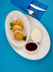 Round cottage cheese pancakes in a white plate on a blue background. Food breakfast. Healthy food. Sour cream sauce and mint. Top view closeup. Dessert meal. Curd cheese pancake ricotta fritters.