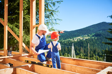 Father with toddler son building wooden frame house. Male builder showing his son the construction...