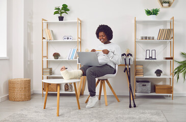 Smiling African American woman with fractured leg talking on video call. Cheerful beautiful girl...