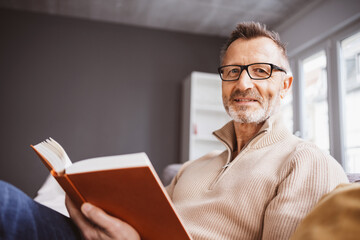 An elderly man with gray hair, a beard and glasses smiles while sitting indoors, casually dressed...