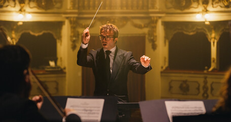 Cinematic Close Up of Conductor Directing Symphony Orchestra with Performers Playing on Stage During Music Concert. Professional Conductor Leading Musicians Passionately in Classic Theater - obrazy, fototapety, plakaty