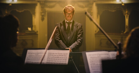 Cinematic Portrait of Male Conductor Standing on Stage Under Spotlight Infront of Symphony Orchestra Musicians. Maestro Focusing and Preparing to Direct a Big Music Concert in Classical Theatre  © Kitreel