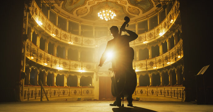 Cinematic shot of Male Cellist Playing Cello Solo on an Empty Classic Theatre Stage with Dramatic Lighting. Professional Musician Rehearsing Before the Start of a Big Show. Silhouette Aesthetics