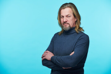 Fototapeta premium Portrait of mature bearded man in turtleneck looking at camera standing with his arms crossed on blue background