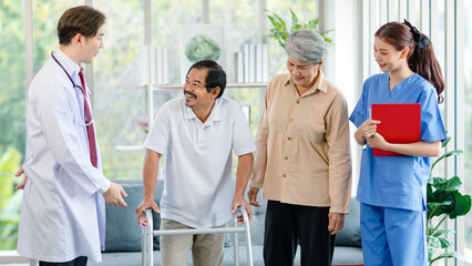 Asian professional successful male doctor in white lab coat with stethoscope female nurse in uniform helping old senior elderly pensioner male husband patient practice walking with four legs walker