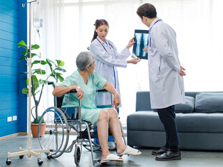 Asian young professional successful male and female doctor in white lab coat with stethoscope standing holding leg bone xray film showing to old senior pensioner injury patient sitting on wheelchair