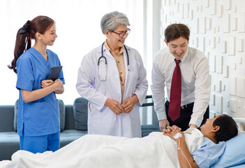Obraz premium Asian senior old professional successful female doctor in white lab coat with stethoscope and internship nurse in blue uniform with insurance staff smiling visiting male patient laying down on bed.