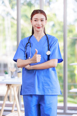 Millennial Asian young beautiful professional successful female internship nurse in blue clinical uniform with stethoscope standing smiling posing crossed arms in hospital office hall way