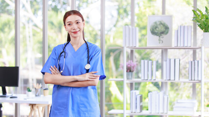 Millennial Asian young beautiful professional successful female internship nurse in blue clinical uniform with stethoscope standing smiling posing crossed arms in hospital office hall way