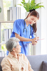 Asian professional successful female intern nurse in blue uniform with stethoscope standing talking asking symptoms from old senior elderly pensioner woman patient and writing note down in clipboard