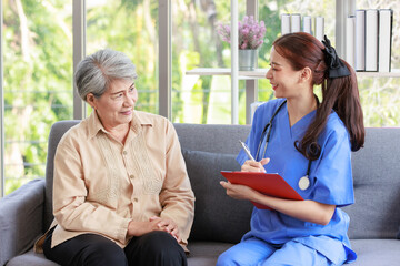 Asian professional successful female intern nurse in blue uniform with stethoscope sitting talking asking symptoms from old senior elderly pensioner woman patient and writing note down in clipboard