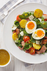Summer fresh salad with potatoes, tomatoes, bacon, arugula, zucchini, cheese and soft-boiled eggs close-up on a plate on the table. Vertical top view from above