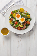 Fresh peasant salad with arugula, spinach, potatoes, bacon, tomatoes, cheese and soft-boiled eggs close-up on a plate on the table. Vertical top view from above