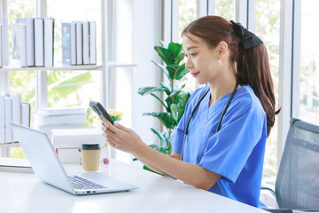 Asian young beautiful professional successful female doctor in blue uniform with stethoscope sitting smiling taking coffee break on call with smartphone in hospital office browsing laptop computer