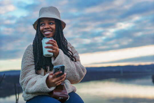 Portrait of a beautiful black woman smiling excitedly at the camera, she's wearing a bucket hat and a backpack, she's drinking coffee and taking pictures with her phone