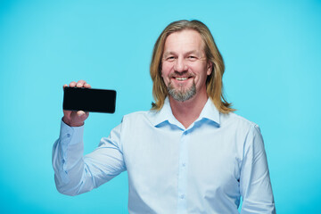 Portrait of successful mature businessman in blue shirt showing his smartphone against blue background