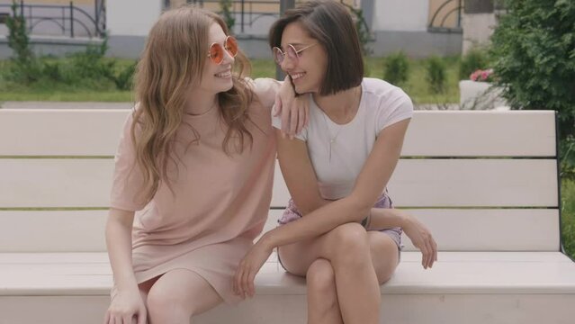 Two young beautiful smiling hipster female in trendy summer clothes.Sexy carefree women sitting on the bench on street background