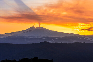 mysterious landscape of great erupting volcano with smoke from craters and snow on slopes in orange light of sunset. eruption of vulcan