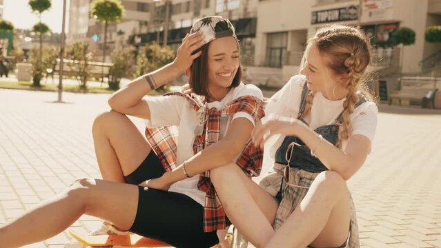 Two young smiling beautiful female with colorful penny skateboards. Women in summer hipster clothes sitting in the street background. Positive models having fun and going crazy