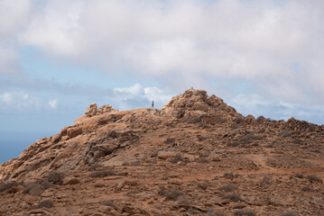 Hombre caminando sobre una ladera rocosa y montañosa en medio de un paisaje desértico en un día soleado con cielo nublado en las Islas Canarias de Fuerteventura.