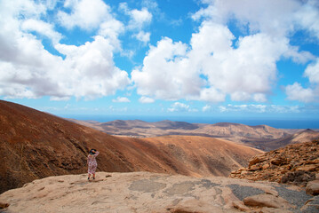 Fototapeta na wymiar Vista panorámica de una mujer rubia con sombrero de paja en medio de un impresionante paisaje volcánico y desértico, Mirador de las Peñas en Fuerteventura, Islas Canarias durante un día soleado.