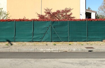 House fence made of net and green tarpaulin. Plants and wall on behind, concrete tile sidewalk and...