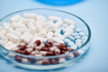 Selective focus on medical pills and gel translucent capsules with granules inside it scattered in petri dish over isolated blue background. World Health Day. Pharmaceutical business and industry