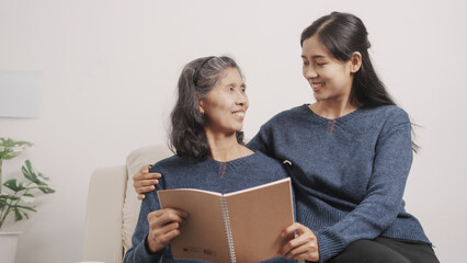Fototapeta na wymiar Reading a book, Mother's day concept, young adult female daughter congratulate excited asian elderly mother at sofa with birthday anniversary, two generations family photo, real people.