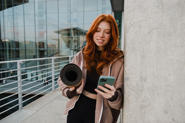 Smiling girl using smartphone and holding yoga mat while standing outdoors