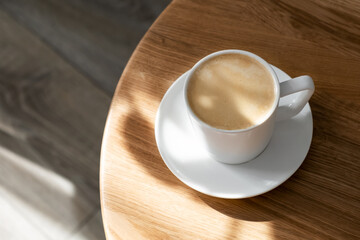 A white porcelain cup with coffee on a saucer stands on a small round table made of natural wood....
