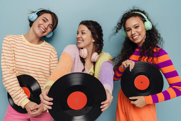 Three beautiful girls in headphones posing with vinyl records and smiling isolated over blue background