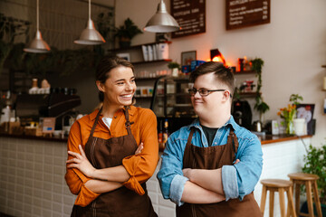 Man with down syndrome and his female colleague smiling while standing in cafe with arms folded