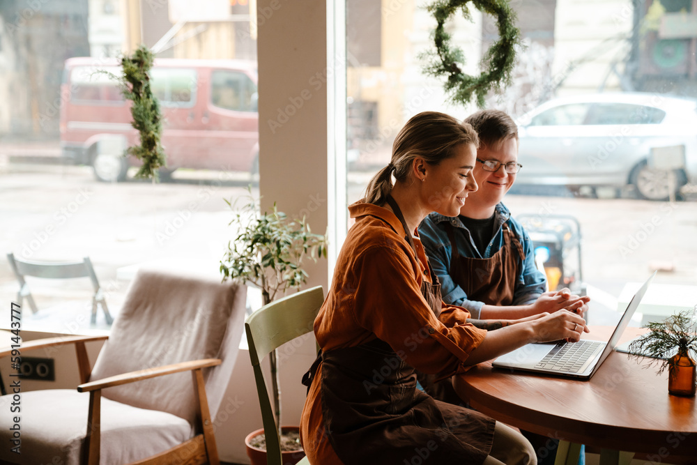 Wall mural Young woman using laptop while training man with down syndrome to work in cafe