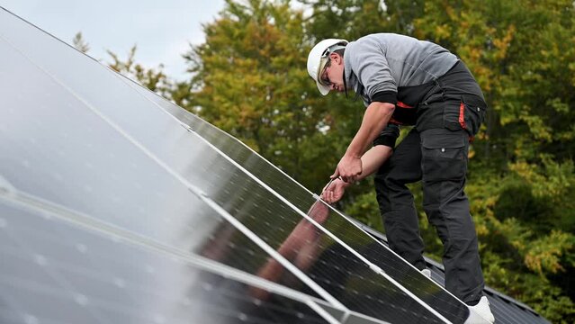 Man technician mounting photovoltaic solar panels on roof of house. Engineer in helmet installing solar module system with help of hex key. Concept of alternative, renewable energy.