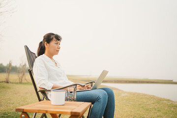 Asian woman typing on laptop while sitting at campsite outdoor. 