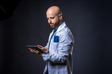 Photo of caucasian serious doctor with beard holding a digital device with blue background and medical white coat. Medical professional using a social app on tablet