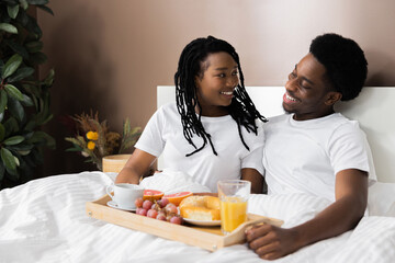 Beautiful adorable African American girl lady talking laughing with boyfriend in bed holding tray with tasty delicious breakfast couple resting at weekends sleeping late.