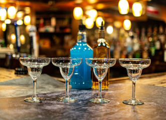 Four Margaritas glasses in front of a blue margaritas mix and a bottle of tequila. The bar in the...