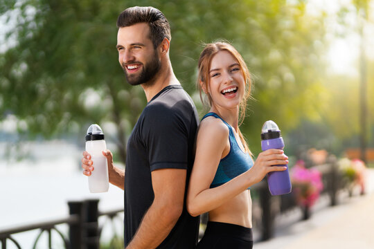 Portrait image of happy smiling, excited couple holding plastic water bottles, woman with man or bearded coach trainer, after successful training, outdoors. Fitness, sport, workout concept.