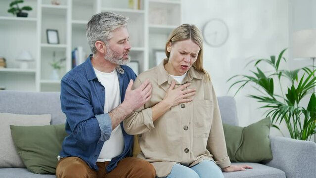 Sick mature woman feels tension pain in chest while sitting on sofa in living room at home. The husband supports and reassures his wife. The female is breathing hard and having a heart attack