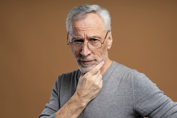 Closeup portrait of 60 years old, serious senior man wearing eyeglasses looking at camera isolated on brown background, holding hand near face. Vision concept