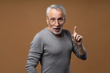 Portrait of excited senior man with open mouth, wearing eyeglasses holding index finger up, having idea isolated on brown background