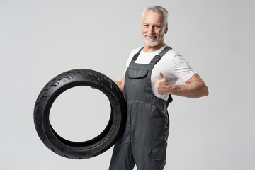 Handsome smiling senior man, mechanic holding seasonal motorcycle tires, showing thumb up wearing overalls isolated on gray background. Successful business, service concept