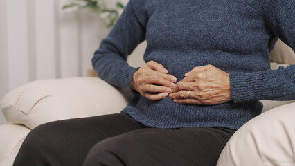 Asian mature woman holding her stomach, grimacing in pain. sitting on sofa.