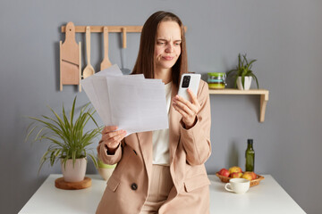 Displeased young woman working out finances in kitchen holding paper document using cell phone, having confused puzzled expression, wearing beige suit.
