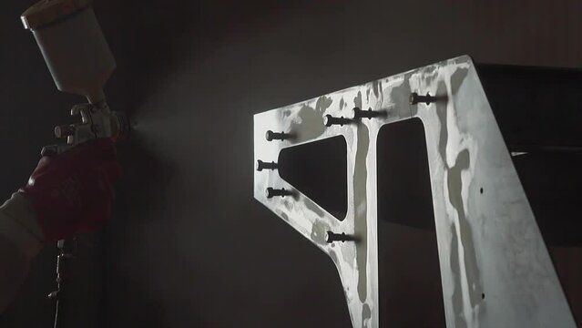 Employee covers metal detail with paint using air brush in workshop closeup. Worker applies color pigment on part with screws at furniture factory